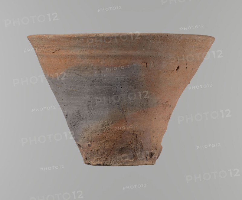 Bowl. Vessel flower pot with a conical shape and a flat bottom. Irregular walls. Straight, flat, slightly flaring, asymmetrical rim. Made on the wheel of Nile clay type NB2 Vienna System. Irregular bottom, cut off with a string; 3.2 cm diameter hole in the bottom. Potter's fingerprints as clear indentations in the lower part next to the bottom. Smooth surface. Reddish-brown color. Condition good, with only minor damage to the clay on the rim. ca. 1550–ca. 1069 BC; New Kingdom, Deir el-Medina. Vessel; height 18 cm, diameter 15.8 cm rim, diameter 9.5 cm bottom., A conical clay pot viewed from the side with a wide rim tapering to a narrower base, showing a rough, textured surface with visible cracks and small pits; the pot displays a mix of reddish-brown and gray areas, with some darker and lighter patches, and a matte, weathered finish against a neutral gray background.