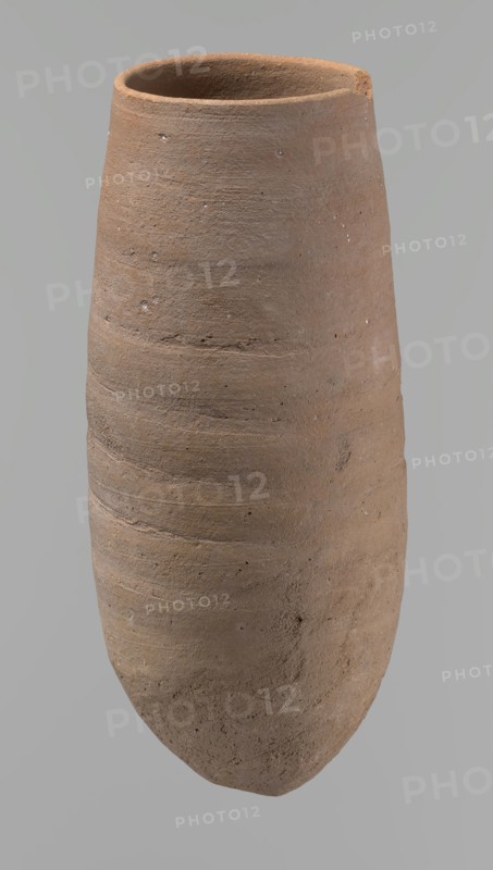 Vessel, Vessel with a conical shape and a pointed bottom. Straight, tapering rim. Made on the wheel of Nile clay type NB2 Vienna System classification. Bottom and outer surface worked with a hard tool. Surface even. Light brown. Fairly well preserved, with minor damage on the rim. c. 1550–c. 1069 BC, New Kingdom, Deir el-Medina. Vessel; height 20.8 cm; diameter 7 cm lip, 9 cm body., A tall, narrow ceramic vessel with a rounded base and an open rim, shown against a plain gray background; the vessel has a textured, slightly rough surface with horizontal ridges and small pits, and its color ranges in shades of brown and tan with subtle darker and lighter variations.