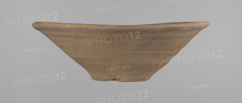 Bowl, Bowl with a conical shape and a flat bottom, slightly asymmetrical. Straight, flaring rim. Vessel made on the wheel of Nile clay type NB2 according to the Vienna System classification. The bottom is cut off with a knife. Surface leveled. The outer part of the vessel is finished to approximately half its height with a knife or similar tool. Color of the vessel: yellow-red. Vessel reconstructed from two original fragments. c. 1550–c. 1069 BC; New Kingdom Ramesside period?, Deir el-Medina. Vessel. Height 9.2 cm; diameter 27.8 cm rim; diameter 8.4 cm bottom., A fragment of a shallow, wide-rimmed ceramic dish in shades of brown and tan is shown against a neutral gray background; the piece has a curved rim with a slightly raised edge and a textured surface with visible horizontal striations and small imperfections, and the fragment tapers toward a narrower base with irregular, worn edges.
