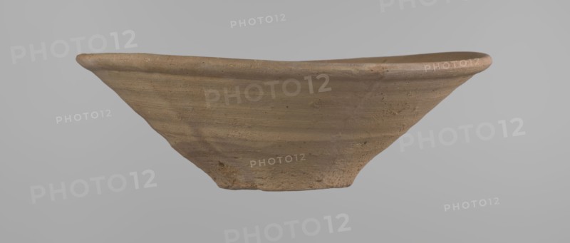 Bowl, Bowl with a conical shape and a flat bottom, slightly asymmetrical. Straight, flaring rim. Vessel made on the wheel of Nile clay type NB2 Vienna System. The bottom is cut off with a knife. Surface leveled. The outer part of the vessel is finished to approximately half its height with a knife or similar tool. Color of the vessel: yellow-red. Vessel reconstructed from two original fragments. c. 1550–c. 1069 BC; New Kingdom Ramesside period?. From Deir el-Medina. Ceramic bowl; height 9.2 cm, rim diameter 27.8 cm, bottom diameter 8.4 cm., A shallow, wide bowl-shaped object made of a coarse, clay-like material in shades of tan and light brown, shown from the side against a plain light gray background; the rim is slightly uneven and rounded, the surface displays horizontal wheel-like ridges and subtle texture variations with small rough patches and specks, and the base tapers inward to a narrower, rougher foot.