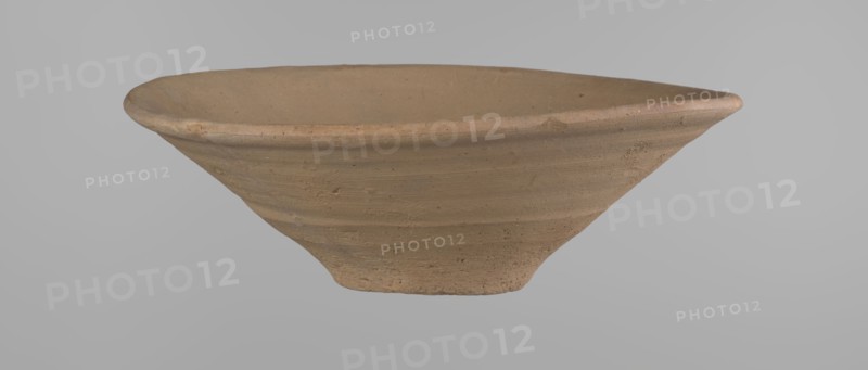 Bowl, Bowl with a conical shape and a flat bottom, slightly asymmetrical; straight, flaring rim. Vessel made on the wheel of Nile clay type NB2 Vienna System. Bottom cut off with a knife; surface leveled. Outer part finished to approximately half its height with a knife or similar tool. Color yellow-red. Vessel reconstructed from two original fragments. c. 1550–c. 1069 BC, New Kingdom Ramesside period?, from Deir el-Medina. Ceramic clay vessel; height 9.2 cm, rim diameter 27.8 cm, bottom diameter 8.4 cm., A shallow, wide bowl-shaped ceramic object with a rough, unglazed surface in shades of tan and light brown, showing concentric horizontal ridges and subtle speckling; the rim is slightly thicker and rounded, while the base tapers inward to a narrower foot; the background is a smooth, uniform light gray.