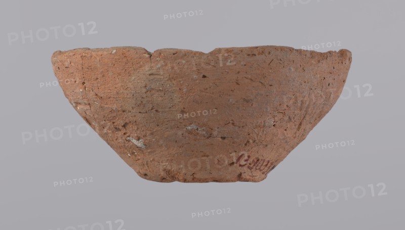 Bowl, probably votive, with a conical shape and a flat bottom. Straight rim, slightly curled towards the center. Wheel-made vessel of Nile clay type NC according to the Vienna System classification. Leveled surface. Flat, irregular bottom, cut off with a string. Vessel color: brown-red. Vessel reconstructed from three original fragments. Rim slightly damaged. c. 1550–c. 1069 BC; New Kingdom, Deir el-Medina. Vessel, height 4.8 cm, diameter 10.2 cm lip, diameter 4 cm bottom, A roughly textured, shallow bowl-shaped object sits against a pale gray background; the object has an uneven rim and a coarse surface with visible pits and small inclusions, showing a dominant reddish-brown clay color with patches of lighter beige and small flecks of gray and white, and a few areas of darker brown; the shape tapers toward the base and the rim edge is irregular and worn.