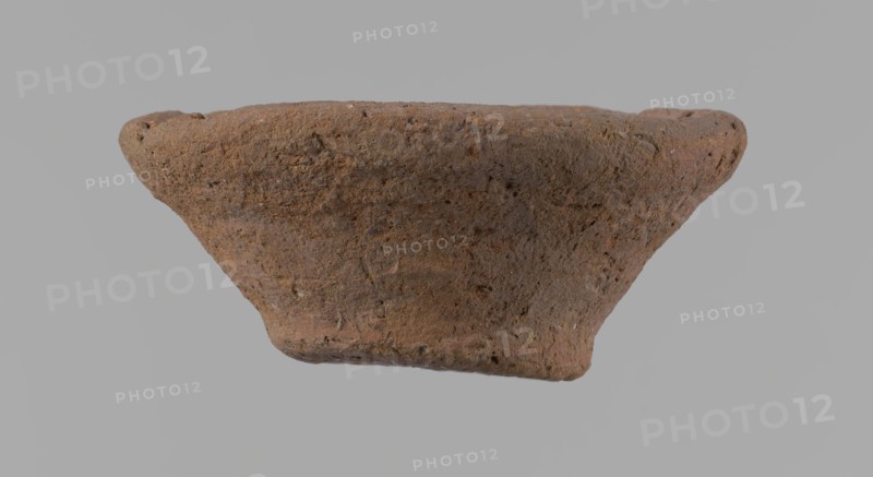 Bowl, probably votive, conical shape with flat bottom. Irregular walls; straight, slightly flaring rim; irregular bottom, probably cut off with a string. Wheel-made vessel of Nile clay type NC Vienna System. Surface only slightly leveled. Vessel color: brown. Well-preserved with only minor surface erosion. c. 1550–c. 1069 BC, New Kingdom, Deir el-Medina. Ceramic bowl, height 3.7 cm; diameter 8.2 cm lip, 4.2 cm bottom., A roughly conical, textured object with an irregular, worn surface fills the center of the image, showing a coarse, grainy texture and pitted areas; the object displays shades of brown and reddish-brown with small darker and lighter speckles, set against a uniform light gray background.