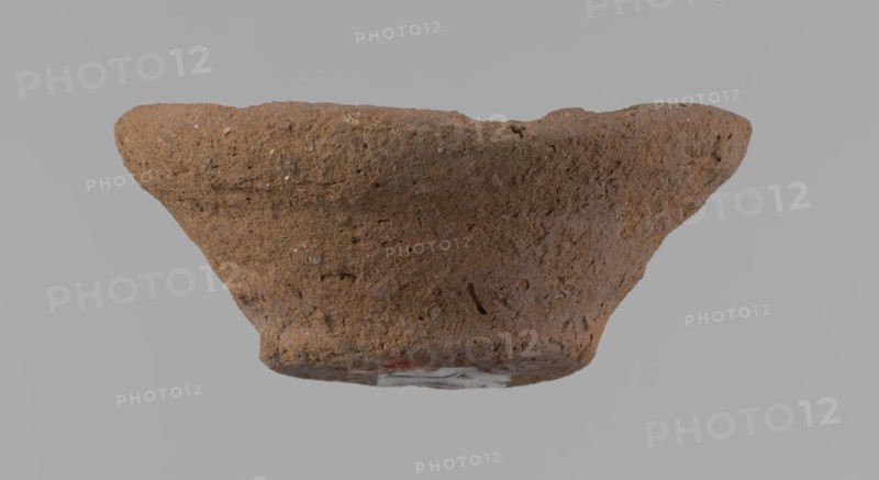 Bowl, probably votive, conical shape with flat bottom. Irregular walls; straight, slightly flaring rim; irregular bottom, probably cut off with a string. Wheel-made vessel of Nile clay type NC Vienna System. Surface only slightly leveled. Vessel color: brown. Well-preserved with minor surface erosion. c. 1550–c. 1069 BC New Kingdom, Deir el-Medina. Vessel; height 3.7 cm; diameter 8.2 cm lip; diameter 4.2 cm bottom., A rough textured, shallow bowl-shaped object with an irregular rim, shown against a gray background; the surface is coarse and porous with small pits and inclusions, and the visible colors are various shades of brown with a few darker brown flecks and a small area of muted red near the base.