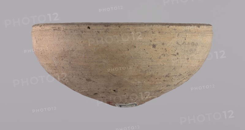 Bowl, Bowl with a conical shape and a pointed bottom. Flat, straight rim. Vessel made on the wheel of Nile clay type NB2 according to the Vienna System classification. Surface even, irregularly covered with engobe inside and outside. Color of vessel: cream. Vessel fairly well preserved, slightly damaged in the upper part. c. 1550–c. 1069 BC; New Kingdom, Deir el-Medina. Vessel; height 6.5 cm, diameter 13.6 cm lip., A large rounded vessel shown from the side and slightly upside down, with a textured surface that appears rough and worn; the vessel is mainly beige with areas of tan and light brown, and it has small darker brown speckles and patches across its surface, a slightly darker rim, and a small area of muted red and a small patch of blue-gray near the bottom; the background is a uniform pale gray.