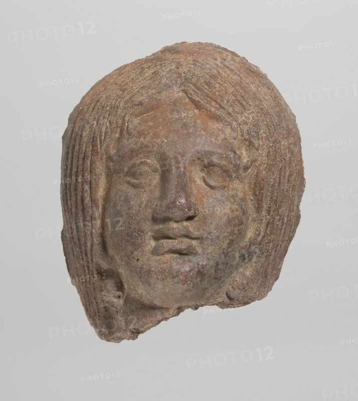 Female votive head, Female head with long, straight hair falling to the shoulders. Neck partially preserved. Elongated face with a receding forehead is characterized by wide-open eyes set shallowly under prominent eyebrow arches. The mouth is slightly open with fleshy lips, the chin protrudes forward, and full cheeks. 2nd century BC, Etruria; votive. Height 20 cm, width 12 cm, depth 8.5 cm, weight 0.94 kg., A worn sculpted head with textured hair and a neutral facial expression, rendered in earthy tones of brown and reddish-brown with patches of darker and lighter brown and some grayish areas; the hair is styled in layered, ribbed strands framing the face, the eyes are almond-shaped and recessed, the nose is rounded, and the lips are full and slightly parted; the surface shows roughness and pitting and fragments missing along the lower edge, set against a pale, light gray background.