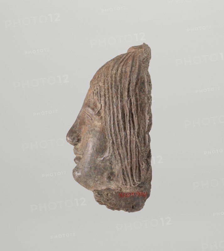 Female votive head, Female head with long, straight hair falling to the shoulders. Neck partially preserved. Elongated face with a receding forehead characterized by wide-open eyes set shallowly under prominent eyebrow arches. Mouth slightly open with fleshy lips, chin protruding forward, and full cheeks. 2nd century BC, Etruria, votive. Height 20 cm, width 12 cm, depth 8.5 cm, weight 0.94 kg., A carved stone fragment showing a profile of a human face viewed from the side with closed eye, a prominent nose, and full lips, attached to a larger irregular base; long textured hair with vertical grooves falls down the back of the head. The object displays a range of browns and grayish-browns with some darker and lighter mottling, and the background is a pale gray.