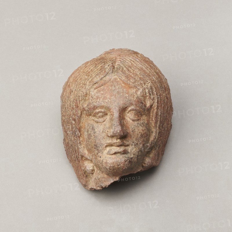 Female votive head, Female head with long, straight hair falling to the shoulders. Neck partially preserved. Elongated face with a receding forehead characterized by wide-open eyes set shallowly under prominent eyebrow arches. Mouth slightly open with fleshy lips, protruding chin, and full cheeks. 2nd century BC, Etruria; votive. Height 20 cm, width 12 cm, depth 8.5 cm, weight 0.94 kg., A weathered sculpted head with textured hair and facial features sits against a plain background; the object shows shades of brown, reddish-brown, tan, and hints of gray, with rough surface variations and small areas of lighter and darker discoloration, and a neutral light gray backdrop.