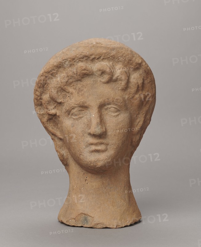 Female votive head, Female head in a wreath, slightly tilted to the right shoulder. Under the wreath are visible regularly arranged locks of hair, framing a low, triangular forehead and temples. Ears are exposed. Deep-set eyes, almost flat in the elaborately worked eyelids, small nose, small mouth with fleshy lips, short, strongly defined chin, full cheeks. 3rd century BC, Etruria. Votive. Height 26 cm, width 16.5 cm, depth 13.5 cm, weight 2.1 kg., A single sculpted head made of a rough, warm brown material sits against a smooth neutral gray background, showing a rounded hairstyle with textured curls, a defined forehead, closed or gently downcast eyes, a straight nose, and a subtle mouth, all with a weathered, pitted surface and small pale flecks; the neck is cylindrical and slightly flared at the base, and the overall tones include warm brown, light tan, small pale spots, and the surrounding gray.