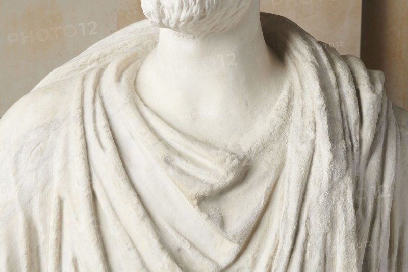 Roman statue, unknown Roman workshop. The figure of a standing man dressed in a tunic and toga, depicted in contrapposto. The right arm is exposed from the elbow, the left is almost completely hidden under the toga. Head from another statue. The face is small, with short stubble laid flat but carefully modeled. The mouth is compact, the corners turned slightly downward. The eyes are large, shadowed by drawn-down eyebrows, with a marked iris and pupil. Rectangular base, unprofiled. 1st–3rd century torso ca. mid-1st century AD; head 3rd quarter of 3rd century AD. Roman Empire 27 BC–476. Sculpture. Weight 870 kg., The image shows a close-up of a sculpture's upper torso and neck draped in fabric-like folds. The surface is predominantly white with subtle off-white and beige tones, and the texture varies from smooth areas on the neck to rougher, carved details on the draped sections. A trimmed beard is visible at the base of the chin, and the drapery forms layered, flowing folds across the c...