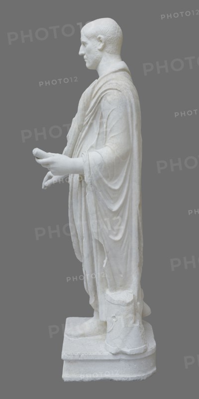Roman statue, unknown Roman workshop. Figure of a striding man dressed in a tunic and toga. The folds of the toga flow widely, with a slit at the left leg. The head is a large addition, with closely cropped hair and beard carved with short cuts of a pointed chisel. The face, with prominent jaws, tight mouth, and drawn eyebrows, has a stern expression. The pupils are marked. Profiled base, rectangular at the front, behind the back of the figure with rounded side edges. Date: 2nd–3rd century torso 2nd century AD, head late 3rd century AD; earlier dating: 1st–2nd century AD. Roman Empire 27 BC–476. Sculpture. Weight 928 kg., A white marble statue of a robed male figure shown in profile against a solid gray background, with the figure standing on a rectangular base and wearing long, draped garments that form several folds and layers; the figure’s head is turned slightly forward, the hair is short and textured, the face shows a calm expression, and both arms are extended forward with hands ...
