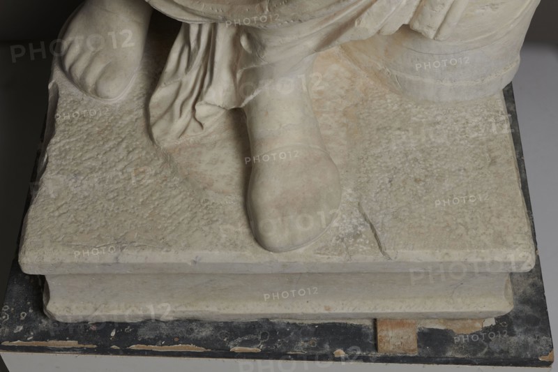 Roman statue, unknown Roman workshop. Figure of a striding man dressed in a tunic and toga. The folds of the toga flow widely, a slit at the left leg. The head is a large addition, with closely cropped hair and beard, carved with short cuts of a pointed chisel. The face, with prominent jaws, tight mouth, and drawn eyebrows, is characterized by a stern expression. The pupils are marked. Profiled base, rectangular at the front, behind the back of the figure with rounded side edges. 2nd–3rd century torso 2nd century AD, head late 3rd century AD; earlier dating 1st–2nd century AD. Roman Empire 27 BC–476. Sculpture. Weight 928 kg., The image shows the lower portion of a sculpture resting on a rectangular stone base, including a carved foot and draped fabric. The stone surfaces display a textured, worn appearance with subtle variations of off-white, beige, and pale gray, and faint patches of light pinkish tones. The base sits on a dark, chipped platform with areas of black, brown, and expose...