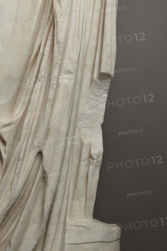 Roman statue, unknown Roman workshop. Figure of a striding man dressed in a tunic and toga. The folds of the toga flow widely, with a slit at the left leg. The head is a large addition, with closely cropped hair and beard carved with short cuts of a pointed chisel. The face, with prominent jaws, tight mouth, and drawn eyebrows, is characterized by a stern expression. The pupils are marked. Profiled base, rectangular at the front, with rounded side edges behind the back of the figure. 2nd–3rd century torso 2nd century AD; head late 3rd century AD; earlier dating 1st–2nd century AD. Roman Empire 27 BC–476. Sculpture. Weight 928 kg., The image shows a close-up of a carved stone surface with draped folds resembling fabric, displaying a smooth pale off-white to cream color with subtle warm beige and light brown discolorations and some darker grayish smudges; the carved folds create deep shadows and rounded ridges, and the right edge transitions to a rougher, broken texture with jagged protr...