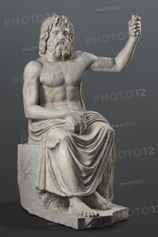 Statue of Jupiter of the Capitoline type, unknown Roman workshop. Jupiter Zeus seated on a throne set on a rectangular base. Seated stiffly with torso slightly reclined; raised left hand holds a scepter, right hand rests on right knee, right foot slightly forward. Legs covered with a himation that also covers the back and left shoulder. Muscular torso strongly developed at the shoulders with naturalistic modeling. Head disproportionately large; face nearly idealized with a slightly open mouth. Hair parted from the center anastole. Thick beard arranged in strands on the sides and in curls on the chin. Roman copy after a Hellenistic variant of Phidias's statue of Zeus the Olympian. Sculpture, late 2nd–3rd century AD Roman Empire. Weight 930 kg., A marble statue of a seated bearded man with long wavy hair, shown from head to feet, posed with one arm raised and the other resting on his lap; he wears a draped garment over his lower body that creates flowing folds, and his exposed torso show...
