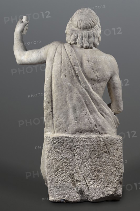 Statue of Jupiter of the Capitoline type, unknown Roman workshop, Scen.Gal.Class., deposit, Collection of Ancient Art, Jupiter mythology, Zeus mythology, Roman sculpture, Roman copy after a Hellenistic variant of Phidias's statue of Zeus the Olympian. Zeus seated on a throne set on a rectangular base. Zeus sits stiffly with his torso slightly back. His raised left hand holds a scepter. His right hand rests on his right knee. The right foot is slightly forward. The legs are covered with a himation, which also covers the back and left shoulder. The muscular torso, strongly developed at the shoulders, has naturalistic modeling. The head is disproportionately large, and the face is perfect with a slightly open mouth. The hair is parted from the center anastole. The beard is thick, arranged in strands on the sides and in curls on the chin. Late 2nd–3rd century, Roman Empire 27 BC–476 AD, sculpture, weight 930 kg, A rear view of a stone sculpture depicting a seated male figure with wavy hair...