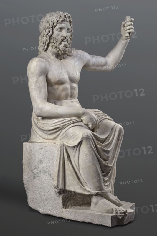Statue of Jupiter of the Capitoline type, unknown Roman workshop. Jupiter Zeus seated on a throne set on a rectangular base. Seated stiffly with torso slightly reclined; raised left hand holds a scepter, right hand rests on right knee, right foot slightly forward. Legs covered with a himation that also covers the back and left shoulder. Muscular torso, strongly developed shoulders, naturalistic modeling. Head disproportionately large; face with slightly open mouth. Hair parted from the center anastole. Beard thick, arranged in strands on the sides and in curls on the chin. Roman sculpture, Roman copy after a Hellenistic variant of Phidias's statue of Zeus the Olympian. Late 2nd–3rd century AD Roman Empire. Sculpture, weight 930 kg., A seated marble statue of a bearded man with long wavy hair, shown shirtless with a draped cloth over his lower body, sitting on a block-like seat; his right arm is raised and bent at the elbow while his left arm rests across his lap, and his feet are bare ...