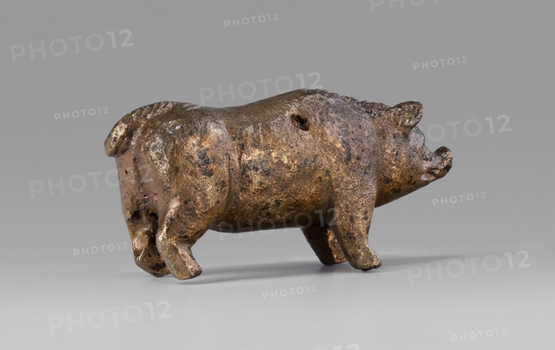 A small bronze figurine of a pig. The animal stands on rather schematically depicted legs ending in forward-pointing hooves. The animal's massive body is characterized by an unnatural length. The torso is adorned with a small, doubled tail. The presence of hair is clearly marked on the animal's back. The pig's muzzle, set on a thick neck, ends in a narrow snout. The pig's slightly protruding ears point forward. The animal's eyes, which are unusually large relative to the rest of the body, are clearly marked. The sculpture is covered with a natural patina. The figurine was probably used as a votive offering; this interpretation is supported by its finding in the vicinity of the temple. Hellenistic period, Kyrene Libya. Figurine. Height 1.8 cm; width 4.2 cm; depth 1.4 cm; weight 0.035 kg., A small sculpted figure of an animal resembling a pig is shown in profile on a smooth surface, with a rounded body, short legs, a curled tail, and a snout; the surface of the figure is textured with ir...
