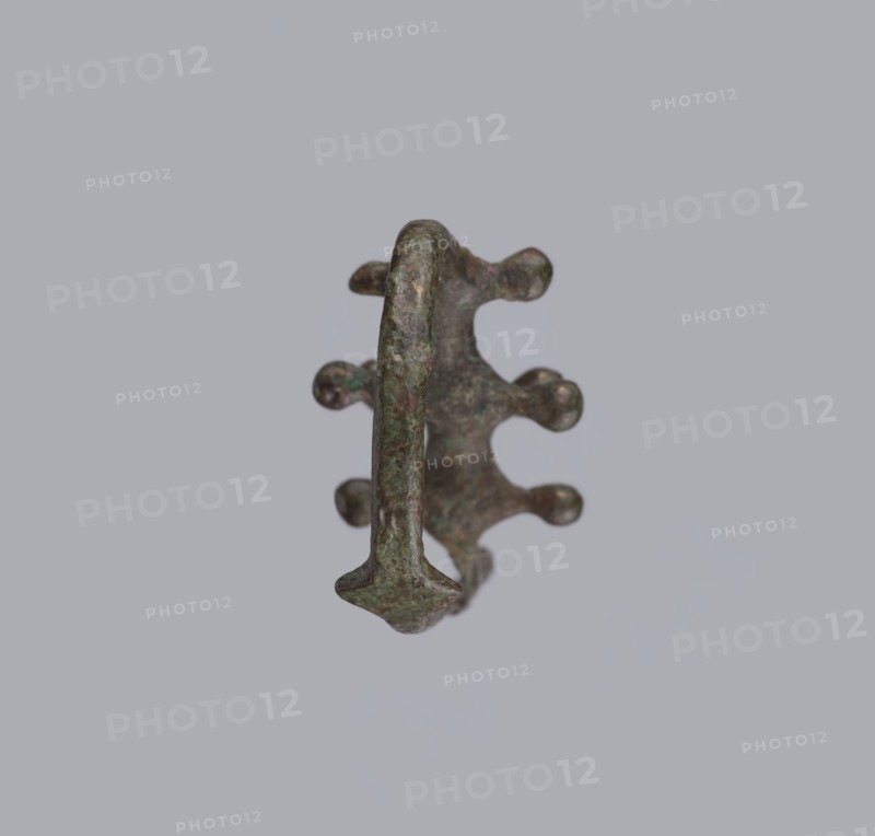 Fibula a Drago, One-piece bronze fibula with an arched ring of circular cross-section bearing four pairs of protrusions terminating in spherical bulges. The transition from the ring to the pin is a flat rhombus. Long cylindrical sheath with a slit for the pin. Preservation: ends of the pin and sheath are broken off. Dated 950–750 BC 4th quarter 8th–7th century BC. Possibly from Cumae. Fibula; height 3.2 cm, width 6.5 cm., A single metallic object with a rough, corroded surface sits against a pale gray background; the object shows a curved central band with several rounded protrusions along one side and a flattened arrowhead-like end, with colors including green, brown, and hints of dark gray.