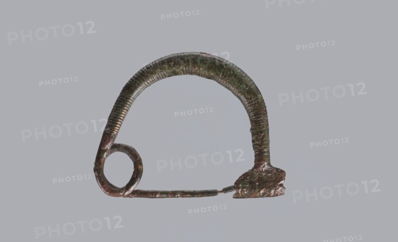 Arched fibula, one-piece ad arco semplice fibula with an arched bow made of a round bronze bar, decorated with groups of circumferential notches. A 1.5-turn loop at the transition from the bow to the pin. Sheath damaged. 9th–8th century BC. Southern Italy, possibly Cumae. Height 3.2 cm, width 4.2 cm., A roughly semicircular metallic object with a textured, ribbed surface and a patina showing green and brown tones, attached on one end to a rounded loop and on the other to an irregular, slightly flattened mount; a thin, straight rod extends between the loop and the mount. The background is a flat, uniform gray.