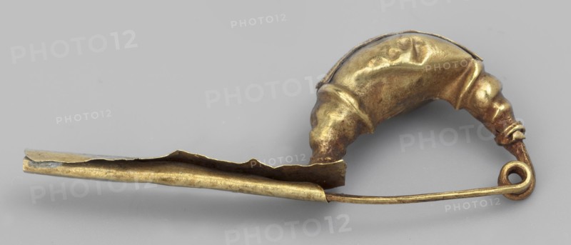 Leech fibula with an elongated stem. The swollen, profiled ring at the ends is decorated with a long strip of filigree in the form of figure eights. The spring has 1 1/2 coils. 2nd half of the 6th century BC, Etruria. Jewelry fibula. Height 1.2 cm; width 3 cm; depth 0.6 cm; weight 1 g., A metallic object photographed on a neutral gray background, showing a curved pin-like form with a looped end and a folded, hollow section; the object has a worn, slightly irregular surface with dents and creases and appears in shades of gold and brass with darker brownish patina in places and small areas of muted gray.
