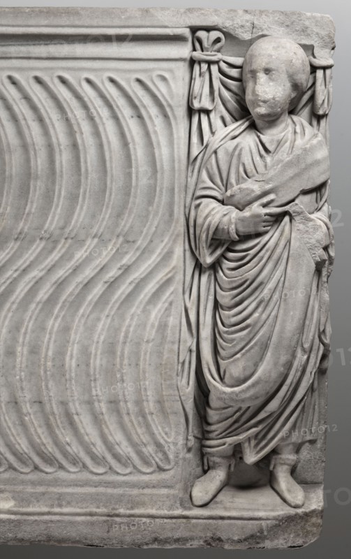 Sarcophagus with a depiction of a married couple and a shepherd, unknown Roman workshop. A large Roman sarcophagus with strigilla S-shaped grooves converging toward the center forming a central medallion, decorated with a standing, thoughtful shepherd leaning on a shepherd's staff pedum. On the front sides are figures presumed to be the deceased, standing before draped fabrics parapetasmata: on the left, a woman in a tunic and long cloak palla holding a scroll volumen in her left hand; on the right, a man in a tunic and toga with contabulatio specially wrapped hem, holding a scroll in his left hand and wearing mullei leather military boots. Both frontal figures have incompletely finished heads. The side walls show reliefs of a seated griffin with a lion's head; the left wall near its lower left corner has an opening into the interior, and the lower left corner of the right wall is missing. The rear wall is rather carelessly worked. Considerable abrasions and damage, particularly at the...