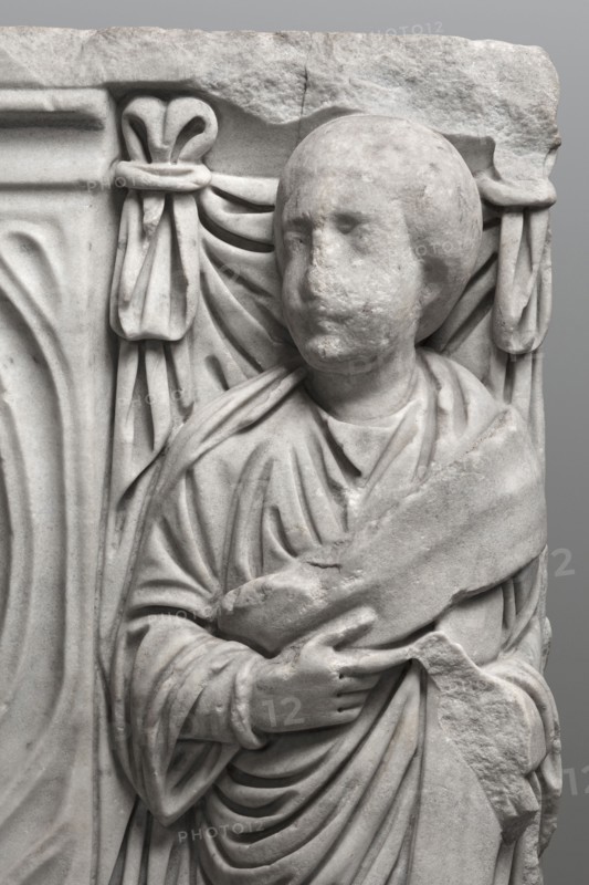 Sarcophagus with a depiction of a married couple and a shepherd, unknown Roman workshop. A large Roman sarcophagus with strigilla S-shaped grooves forming a central medallion, decorated with a standing, thoughtful shepherd leaning on a staff pedum. On the front sides are figures of the presumed deceased standing against draped fabrics parapetasmata: left, a woman in a tunic and long cloak palla holding a scroll volumen in her left hand; right, a man in a tunic and toga with a contabulatio specially wrapped hem holding a scroll in his left hand and wearing mullei leather military boots. Both frontal figures have incompletely finished heads. The side walls show reliefs of a seated griffin with a lion's head; the left side near its lower left corner has an opening to the interior, and the lower left corner of the right wall is missing. The rear wall is more roughly worked. Considerable abrasions and damage, particularly to the corners and the figures on both sides of the front wall. Circa...