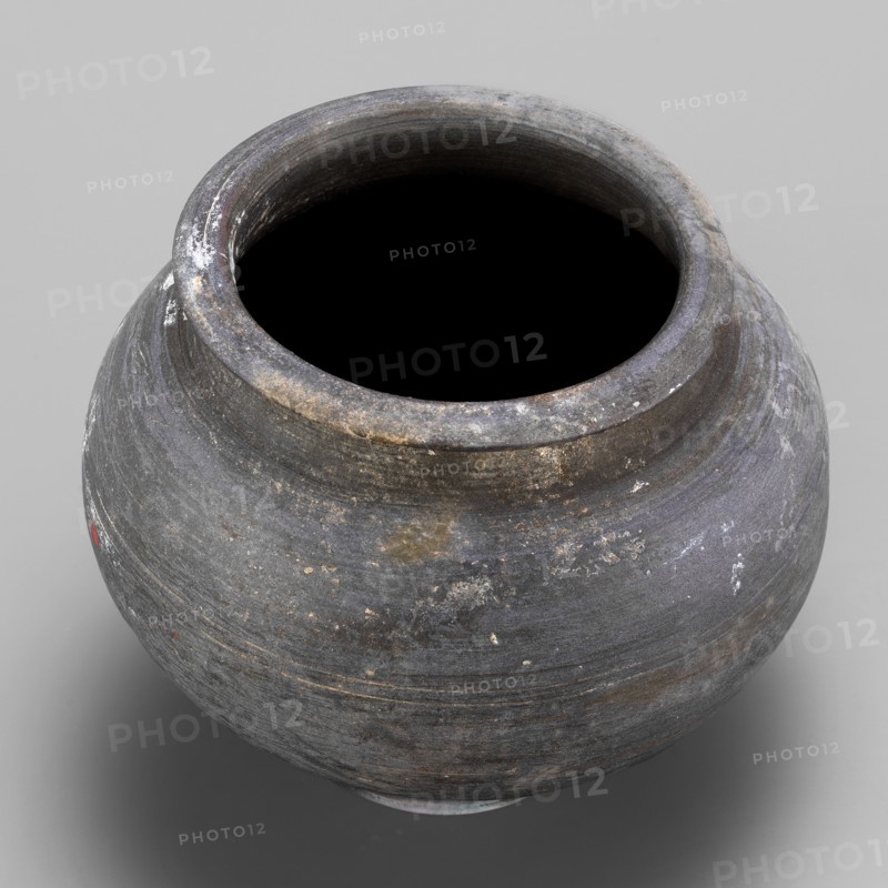 Vessel, unknown northern Mesopotamian workshop; viceless vessel, metallic ware, spherical vessel for oils and ointments; low vessel with a spherical belly, rounded bottom, low wide neck with a narrow, upturned spout. 1st half of the 3rd millennium BC; Early Jazira III period, Northern Mesopotamia Khabur Basin, Tell Rad Shakra Northeastern Syria. Height 8.3 cm, width 10.6 cm, weight 220 g., A rounded ceramic vessel with a wide open mouth sits against a neutral background, showing a weathered surface in shades of gray and charcoal with subtle bluish and brownish tints, areas of lighter whitish residue and small patches of reddish-brown, a few rough spots and fine horizontal striations from its making, and a dark interior visible through the opening.