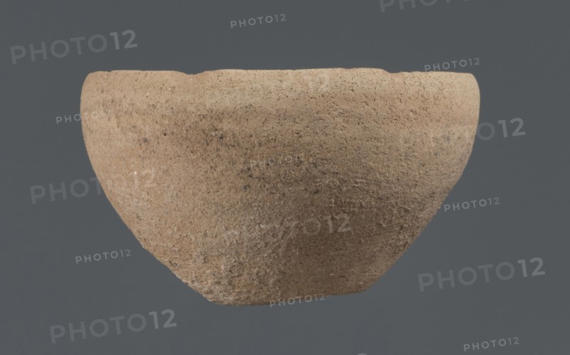 Bowl, unknown Northern Mesopotamian workshop, Early Jazira III period ca. 2600–ca. 2350 BC. Clay vessel tableware. Bowl with a flat, obliquely cut round bottom, gently sloping walls that straighten in the upper part and bend slightly inward, ending in a straight, non-distinctive rim. Before firing, a potter's mark was engraved at the bottom as two intersecting lines of differing lengths: a shorter line parallel to the bottom and a longer line starting at the belly and reaching half the bottom diameter. Origin: Northern Mesopotamia Khabur basin, Tell Rad Shakra, Northeastern Syria., A roughly bowl-shaped object with a coarse, textured surface in shades of tan, beige, and light brown suspended against a flat dark gray background; the rim is uneven and the body shows mottled darker brown speckles and small lighter flecks across its surface.