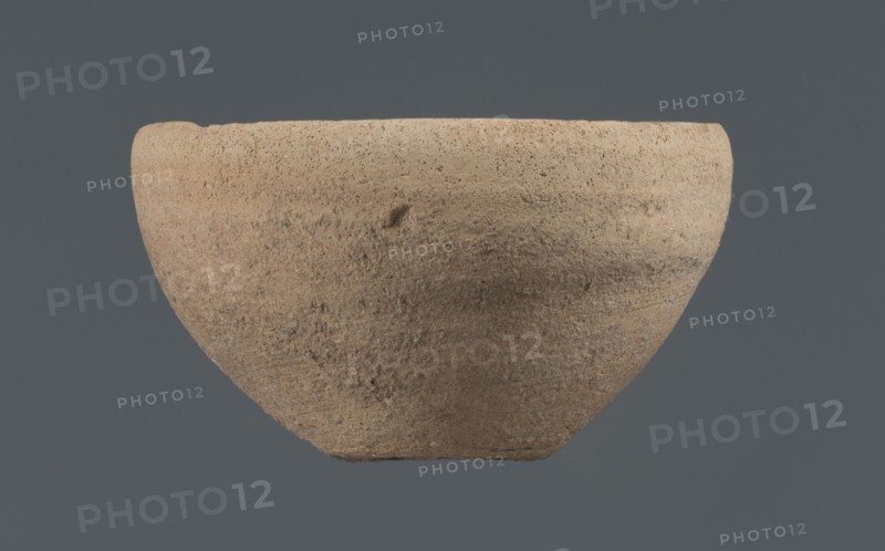 Bowl, unknown Northern Mesopotamian workshop, Early Jazira III period ca. 2600–ca. 2350 BC Early Dynastic III period. Clay vessel: bowl with a flat, obliquely cut, round bottom, gently sloping walls that straighten in the upper part and bend slightly inwards to a straight, non-distinctive rim. Before firing a potter's mark was engraved at the bottom: two intersecting lines of varying lengths — the shorter line runs parallel to the bottom, the longer starts at the belly and reaches half the bottom diameter. Origin: Northern Mesopotamia Khabur basin, Tell Rad Shakra, northeastern Syria., A roughly hemispherical bowl with a coarse, textured surface sits against a solid background; the bowl shows an earthy beige and light brown coloration with subtle darker brown patches and small speckled areas, and the background is a muted grayish blue.