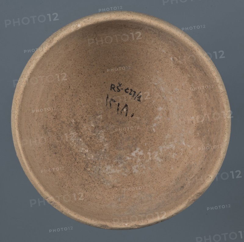 Bowl, unknown Northern Mesopotamian workshop, Early Jazira III period ca. 2600–ca. 2350 BC Early Dynastic III period. Clay vessel: bowl with a flat, obliquely cut round bottom, gently sloping walls that straighten in the upper part and bend slightly inward, ending in a straight, non-distinctive rim. Before firing, a potter's mark was engraved at the bottom as two intersecting lines of varying lengths: a shorter line parallel to the bottom and a longer line starting at the belly and reaching half the bottom diameter. ca. 2600–ca. 2350 BC; northern Mesopotamia Khabur basin, Tell Rad Shakra, northeastern Syria., A round shallow bowl viewed from above with an inner surface showing a rough, sandy texture in shades of light brown and tan with patches of pale whitish residue and darker brown speckling; the rim is slightly thicker and uniform in a beige tone, and the background around the bowl is a muted blue-gray.