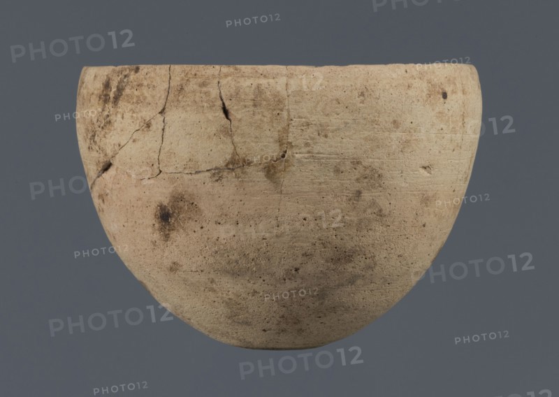 Bowl, unknown northern Mesopotamian workshop, Early Jazira III period ca. 2600–ca. 2350 BC / Early Dynastic III period, clay, bowl with an egg-shaped body, rounded bottom and a straight, non-separated rim slightly bent inwards, ca. 2600–ca. 2350 BC, Northern Mesopotamia Khabur basin, Tell Rad Shakra North-Eastern Syria, Syria; vessel; height 7.8 cm; width 10.7 cm, A light beige rounded clay vessel with a slightly uneven rim and visible surface cracks and repairs sits against a flat muted blue-gray background, showing a textured surface with patches of darker brown discoloration and small pitted marks.