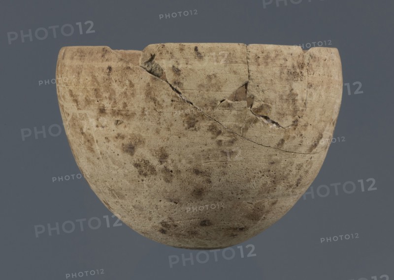 Bowl, unknown northern Mesopotamian workshop, Early Jazira III period ca. 2600–ca. 2350 BC / Early Dynastic III period ca. 2600–ca. 2350 BC. Clay vessel; egg-shaped bowl with a rounded bottom and a straight, non-separated rim slightly bent inward. Northern Mesopotamia Khabur basin; found at Tell Rad Shakra, North-Eastern Syria. Vessel; height 7.8 cm, width 10.7 cm., A rounded ceramic bowl with a chipped rim and visible cracks sits against a solid gray background; the bowl shows a weathered, mottled surface in beige and tan with darker brown spots and smudges, and a few lighter worn areas where the material appears paler.