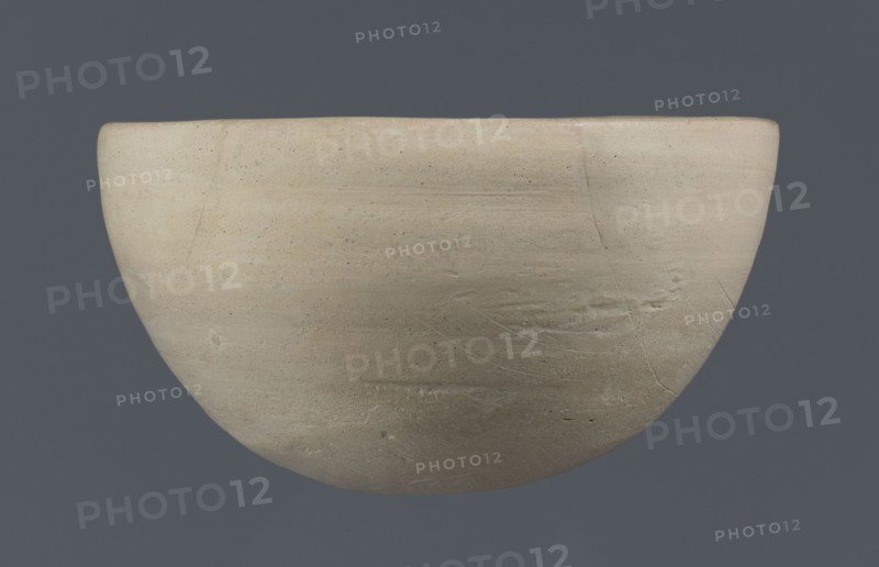 Bowl, unknown Northern Mesopotamian workshop; clay; Early Jazira III period ca. 2600–ca. 2350 BC / Early Dynastic III period. Tableware vessel with a flat, slightly oblique oval bottom with traces of being cut off with a string, gently tilted walls that straighten and bend slightly inwards in the upper part, and a straight, undifferentiated rim. Northern Mesopotamia Khabur basin, Tell Rad Shakra Northeastern Syria., A rounded bowl with a smooth, matte surface in shades of beige and light tan set against a solid dark gray background; the bowl shows subtle horizontal striations, fine surface speckles, and a few shallow scratches and hairline cracks, with slight variations in tone creating areas of warmer cream and cooler gray-beige.