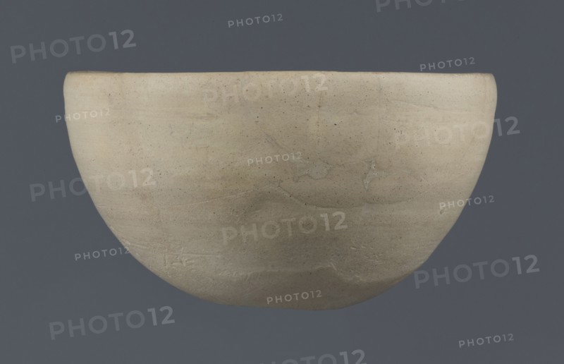 Bowl, unknown Northern Mesopotamian workshop, clay, Early Jazira III period ca. 2600–ca. 2350 BC Early Dynastic III period. Tableware bowl with a flat, slightly oblique, oval bottom with traces of being cut off with a string, gently tilted walls that straighten and bend slightly inwards in the upper part, and a straight, undistinguished rim. Origin: Al-Jazira Khabur basin, Northern Mesopotamia Tell Rad Shakra, northeastern Syria., A smooth rounded bowl with a matte surface suspended against a flat background, the bowl showing variations of beige and light tan with subtle darker speckles and faint irregular streaks, and the background appearing as a uniform muted bluish-gray.