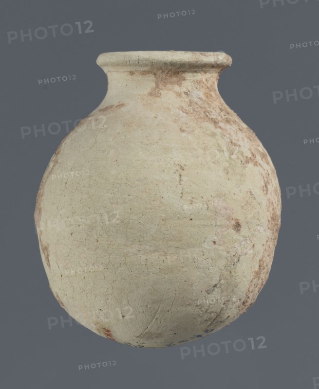 Spherical vessel, unknown northern Mesopotamian workshop, clay, Early Jazira III / Early Dynastic III period ca. 2600–ca. 2350 BC. Spherical vessel with an egg-shaped belly with a rounded bottom and a low cylindrical neck ending with a thickened rim turned outwards. Origin: Northern Mesopotamia Khabur basin, Tell Rad Shakra, Syria. Vessel; height 7.8 cm, width 6.5 cm., A rounded clay vessel with a narrow neck and flared rim sits against a solid gray background; the vessel surface is mostly off-white with areas of pale beige and light brown discoloration, visible weathering and surface cracks, and patches of darker brown and muted reddish-brown along one side, with a small area of faint blue and a touch of red near the lower edge.