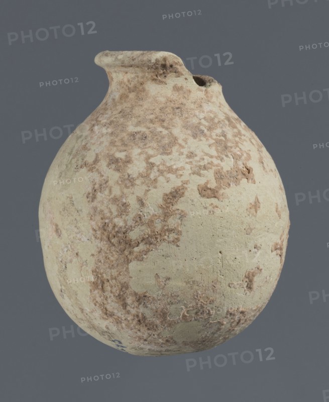 Spherical vessel, unknown northern Mesopotamian workshop, clay, Early Jazira III period ca. 2600–ca. 2350 BC; Early Dynastic III period, Al-Jazira northern Mesopotamia, Khabur basin, Tell Rad Shakra Tell Rad Shakrah?, North-Eastern Syria; spherical vessel with an egg-shaped belly, rounded bottom, low cylindrical neck with thickened outward-turned rim; height 7.8 cm, width 6.5 cm., A rounded ceramic jug with a short neck and a slightly flared rim, shown against a flat gray background; the jug surface is mottled with pale beige and off-white base tones and irregular patches of light to medium brown encrustation, with a rough, weathered texture and small pits and flaking across its body.