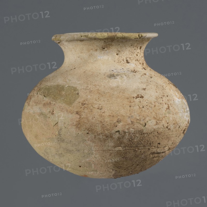 Vessel with an ellipsoidal belly, unknown northern Mesopotamian workshop, Early Jazira III period ca. 2600–ca. 2350 BC / Early Dynastic III period ca. 2600–ca. 2350 BC; clay vessel for oils and ointments; a small vessel with an ellipsoidal belly, rounded bottom, low flared neck ending in a flat top, thickened by an outward curl; Al-Jazira Khabur basin, Northern Mesopotamia Tell Rad Shakra, northeastern Syria; height 7.4 cm, width 8.6 cm., A weathered rounded clay vessel with a narrow neck and flared rim sits against a flat gray background; the pot displays a rough, pitted surface with patches of lighter beige, tan, pale yellow, and muted green, along with areas of brown discoloration and faint whitish deposits, and its texture shows cracks, chips, and uneven wear.