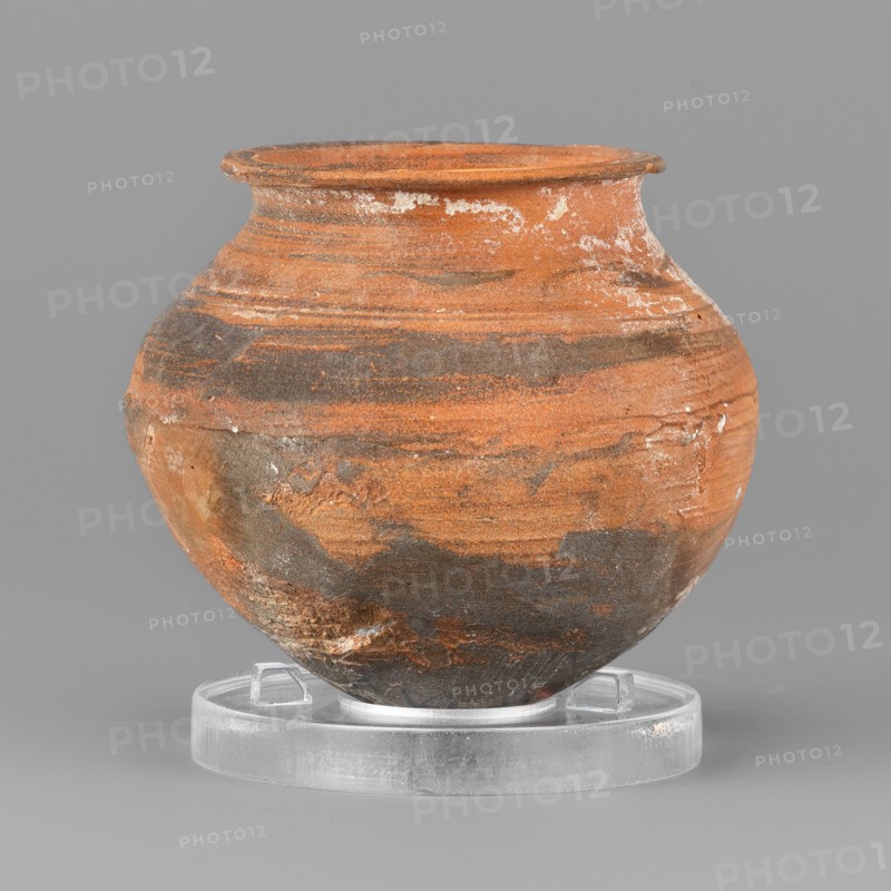 Vessel, unknown northern Mesopotamian workshop, metallic ware; viceless vessel for oils and ointments; clay miniature vessel with an ovoid belly, pointed bottom, low neck terminating in a narrow upturned rim. 1st half of the 3rd millennium BC; Early Jazira III period, Northern Mesopotamia Khabur Basin, Tell Rad Shakra Northeastern Syria. Height 5.5 cm, width 6.1 cm, weight 60 g., A round ceramic pot with a flared rim sits on a clear circular stand; the pot shows a textured surface with a mix of orange and dark gray tones forming layered, streaky patterns, plus small areas of white residue and lighter beige patches, all set against a neutral gray background.