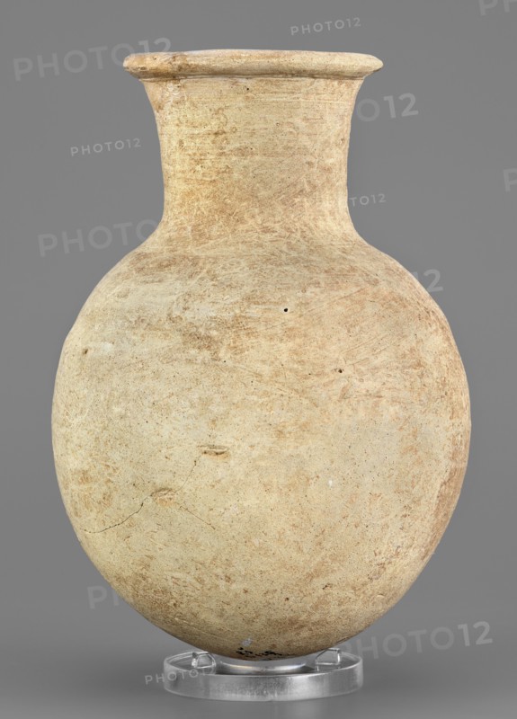 Flask, unknown Northern Mesopotamian workshop. Clay flask-shaped vessel with an elongated ovoid belly, rounded bottom, high slightly conically flaring neck separated from the belly by narrow shoulders and ending with a flat top, protruding externally thickened rim with a triangular cross-section. 2nd half of the 3rd millennium BC; Early Jazira IV Akkadian period, ca. 2350–2200 BC. Northern Mesopotamia Khabur basin, Tell Rad Shakra North-Eastern Syria. Height 14.8 cm, width 10.2 cm, weight 315 g., A rounded vessel with a narrow neck and flared rim sits upright on a clear circular stand; the surface of the vessel shows a textured, worn appearance with patches of lighter and darker beige and tan, along with subtle reddish-brown areas and small darker specks; the background is a flat medium gray.