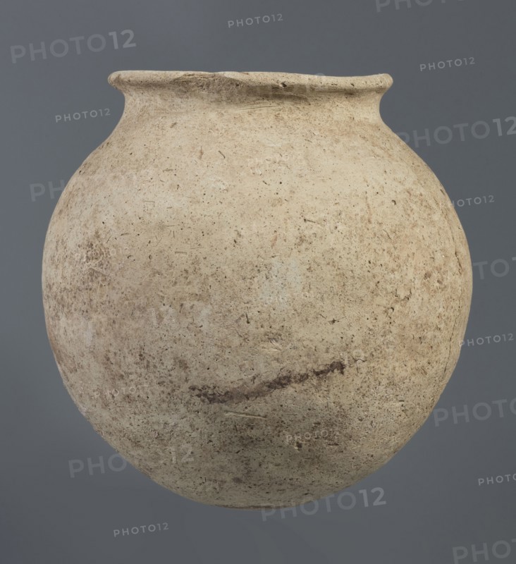 Spherical clay vessel, vice-less, from an unknown Northern Mesopotamian workshop, Early Jazira III / Early Dynastic III period ca. 2600–ca. 2350 BC. Vice-less spherical vessel with rounded bottom; upper wall bends inward forming sloping arms that merge into an arched low neck terminating in a narrow, outwardly obliquely cut rim with a triangular cross-section. Origin: Northern Mesopotamia Al-Jazira, Khabur basin, Tell Rad Shakra, Northeastern Syria. Height 20.3 cm, width 21 cm., A rounded, weathered clay vessel with a narrow flared rim fills the frame against a solid gray background, showing a rough, pitted surface with a palette of beige, tan, cream, light brown, and hints of darker brown; the vessel displays irregular speckling, smudges, and subtle linear marks across its body and rim, with variations in texture from smoother upper areas to more grainy, mottled lower sections.