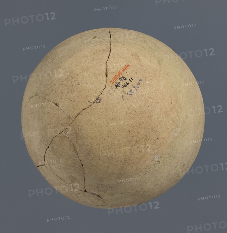 Spherical vessel, unknown Northern Mesopotamian workshop, Early Jazira III period ca. 2600–ca. 2350 BC / Early Dynastic III period ca. 2600–ca. 2350 BC. Spherical vessel with an egg-shaped belly and a rounded bottom; in the upper part the belly merges into a low cylindrical neck terminating in a narrow, strongly protruding, rounded rim turned outwards. Clay, vice-free bulbous vessel. Origin: Al-Jazira Khabur basin, Northern Mesopotamia; Tell Rad Shakra northeastern Syria. Height 16.5 cm, width 16.2 cm., A round, weathered object with a rough, sandy surface in shades of beige and tan, showing a few dark cracks and hairline fractures across its surface; there is a small area with traces of darker markings in muted blue and red, and a few tiny pale spots and scuffs; the background is a solid muted gray.