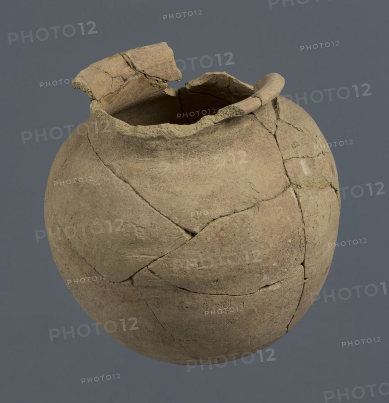 Spherical vessel, unknown Northern Mesopotamian workshop; Early Jazira III period ca. 2600–ca. 2350 BC; Early Dynastic III period ca. 2600–ca. 2350 BC. Spherical vessel with an egg-shaped belly and a rounded bottom. In the upper part the belly merges into a low cylindrical neck terminating in a narrow, strongly protruding, rounded rim turned outwards. Clay vessel; vice-free, bulbous form. Origin: Northern Mesopotamia Khabur basin, Tell Rad Shakra, northeastern Syria. c. 2600–ca. 2350 BC. Height 16.5 cm; width 16.2 cm., A weathered, round pottery vessel with a broken rim and missing fragments showing visible cracks and repaired joins, displaying a rough, uneven surface texture; the vessel is primarily tan and beige with areas of darker brown and traces of lighter off-white, set against a plain gray-blue background.