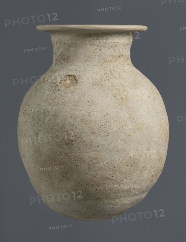 Vise-less jug, unknown northern Mesopotamian workshop. Early Jazira III / Early Dynastic III period, ca. 2600–2350 BC. Clay vessel: vise-less jug with an ovoid belly, flat crookedly cut round bottom, sloping shoulders rising to a high cylindrical neck terminating in a wide flat spout protruding outwards. Bulbous vessel; jug. Origin: Northern Mesopotamia Khabur basin, Al-Jazira. Found at Tell Rad Shakra northeastern Syria., A large rounded clay vessel with a wide flared rim sits against a solid dark gray background. The vessel's surface is a pale beige to off-white with areas of light brown and faint tan discoloration, showing a textured, slightly pitted and weathered finish. There are visible repaired seams and subtle irregularities in shape, including a rough circular patch on one side. The rim is smoother and lighter in tone than the body, and the overall appearance is matte with small speckles and faint mottling of beige, tan, and light brown.