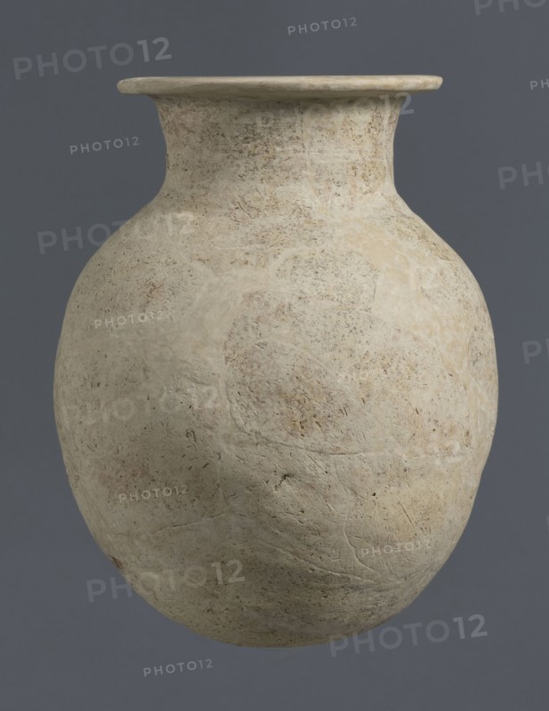Vise-less jug, unknown northern Mesopotamian workshop; Early Jazira III period ca. 2600–ca. 2350 BC; clay; bulbous vessel — jug vise-less. Vise-less jug with an ovoid belly with a flat, crookedly cut round bottom, sloping shoulders, from which arises a high cylindrical neck terminating in a wide flat spout protruding outwards. Origin: Al-Jazira northern Mesopotamia, Khabur basin; discovered at Tell Rad Shakra, northeastern Syria., A large rounded vessel with a narrow neck and flared rim sits against a solid background; the vessel surface appears rough and weathered with an uneven texture and small pitting and scratches, showing a pale beige to off-white color with areas of light tan and faint gray mottling, while the background is a muted dark gray-blue.