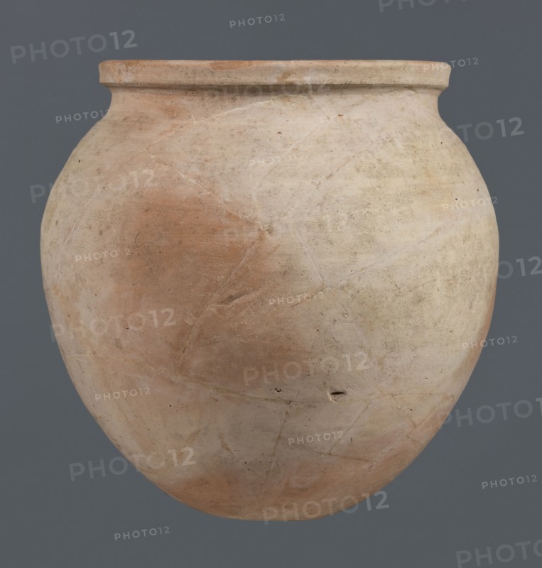 Spherical vessel, unknown northern Mesopotamian workshop, Early Jazira III period ca. 2600–ca. 2350 BC / Early Dynastic III period ca. 2600–ca. 2350 BC. Spherical vessel with an ovoid belly widening towards the top with a small, flat, round bottom, short sloping shoulders merging into a low, wide neck terminating in a rounded rim having the form of a wide, thickened strip on the outside. Clay vessel, vice-free, height 28 cm, width 28 cm. Origin: Northern Mesopotamia Khabur basin, Tell Rad Shakra, Syria northeastern Mesopotamia., A large rounded clay vessel with a short rim, showing a surface of muted beige and pale tan with areas of light reddish-brown and subtle grayish tones, visible texture and fine cracks across its body, small dark specks and a few tiny pits, set against a uniform dark gray background.