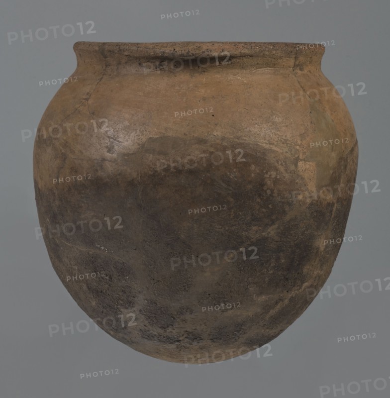 Kitchen pot, unknown northern Mesopotamian workshop, Early Jazira III period ca. 2600–ca. 2350 BC. A kitchen pot with an egg-shaped belly and rounded bottom. Upper walls bend slightly inward forming narrow shoulders that give rise to a low, cylindrical wide neck with a straight, slightly tilted spout. Two slightly thickened horizontal crescent-shaped handles are attached on opposite edges of the spout. Outer walls are intentionally roughened at the bottom up to half the vessel’s height and carefully polished above. Traces of soot are visible on the bottom and slightly above. Syria, Tell Rad Shakra northeastern Syria. Vessel; height 20.5 cm, width 20.7 cm., A large rounded clay pot with a narrow rim in shades of brown and tan, showing darker brown and almost black areas on its lower half and lighter tan patches near the top, with an overall textured, weathered surface and a muted gray background.