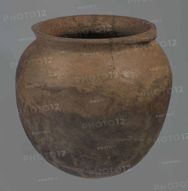 Kitchen pot, unknown northern Mesopotamian workshop, Early Jazira III period ca. 2600–ca. 2350 BC. A kitchen pot with an egg-shaped belly and a rounded bottom. In the upper part the walls bend slightly inwards, forming narrow arms that give rise to a low, cylindrical, wide neck with a straight, slightly tilted spout. Two slightly thickened horizontal crescent-shaped handles are attached on opposite edges. The outer walls are intentionally roughened at the bottom up to half the vessel's height, while above they are carefully polished. Traces of soot are visible on the bottom and slightly above. Syria, Tell Rad Shakra northeastern Syria. Vessel; height 20.5 cm, width 20.7 cm., A rounded clay pot with a wide, slightly flared rim, showing a weathered, rough surface with visible cracks and texture; the pot displays a gradient of earthy colors including brown, tan, ochre, and darker brown to almost black areas, with mottled patches and variations across its surface against a plain gray backg...