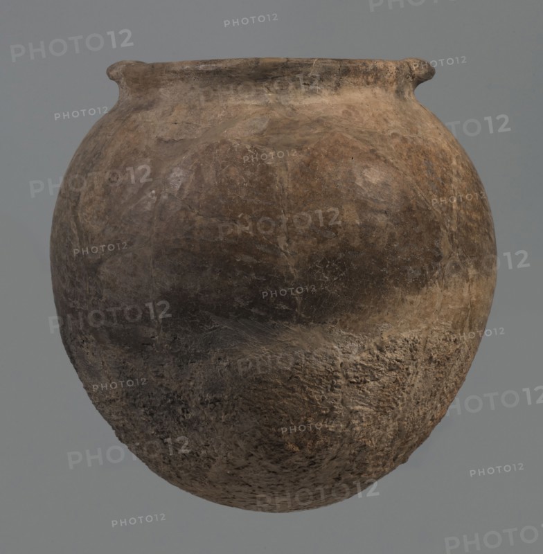 Kitchen pot, unknown northern Mesopotamian workshop. Early Jazira III period ca. 2600–ca. 2350 BC. A kitchen pot with an egg-shaped belly and rounded bottom, terminating in a low, wide cylindrical neck with a rounded, slightly tilted spout, to which two thickened, horizontal crescent-shaped handles are attached on opposite edges. Outer walls intentionally roughened at the bottom up to half the height and polished above. Traces of soot visible at the bottom and slightly above. Fabric contains a rich admixture of crushed quartz. Syria; Tell Rad Shakra northeastern Syria. Northern Mesopotamia place of manufacture. Vessel; height 23.3 cm, width 23.3 cm., A rounded, weathered clay vessel with a narrow, slightly flared rim dominates the image, showing a rough, pitted surface texture and irregular patches of wear; the vessel exhibits a palette of brown tones ranging from light tan to dark brown with areas of gray and muted ochre, and subtle variations in shading that emphasize its uneven, age...