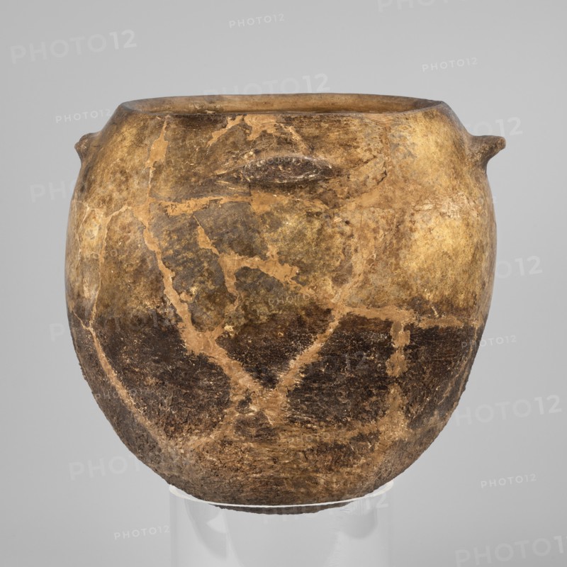 Kitchen pot, unknown northern Mesopotamian workshop, kitchen vessels. A kitchen pot with an egg-shaped belly with a rounded bottom, slightly inward-sloping walls, terminating at the top in a rim, diagonally cut inward. A few centimeters below the rim, four symmetrically placed horizontal crescent-shaped handles are attached. The outer walls are intentionally roughened at the bottom up to 2/3 of the vessel's height, while polished above. The roughened part of the vessel is heavily sooted. The clay contains a rich admixture of crushed quartz. 1st half of the 3rd millennium BC; Early Jazira III period, Tell Rad Shakra northeastern Syria, Northern Mesopotamia. Vessel; height 23.5 cm, width 25.3 cm, weight 2.6 kg., An aged rounded vessel with a wide opening and a slightly flared rim sits against a neutral background, showing a surface mottled with shades of brown, tan, beige, and dark brown; its exterior displays areas of lighter beige patches and darker brown sections with visible irregula...