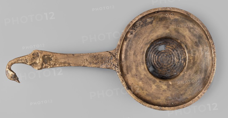 Strainer with a handle in the form of a swan's head;, swans iconography; tools. Wide, flattened container with a hemispherical strainer and a flat handle ending in a bird's head. 2nd half of the 4th century BC previously dated 4th century BC. Etruria. Vessel. Height 23.1 cm; width 11.3 cm; depth 4 cm; weight 0.14 kg., A single metal object resembling a shallow bowl with a long handle is shown against a plain gray background, the object displaying a weathered metallic surface in shades of gold, bronze, brown, and darker tarnished areas; the bowl has a circular recessed center with a patterned perforated disk that appears darker with black and deep brown tones, and the rim and interior show mottled patches of lighter and darker bronze; the handle extends outward with an irregular hooked end and a textured, pitted surface in bronze and gold hues, and the overall piece has an aged, uneven finish with small spots of darker discoloration.